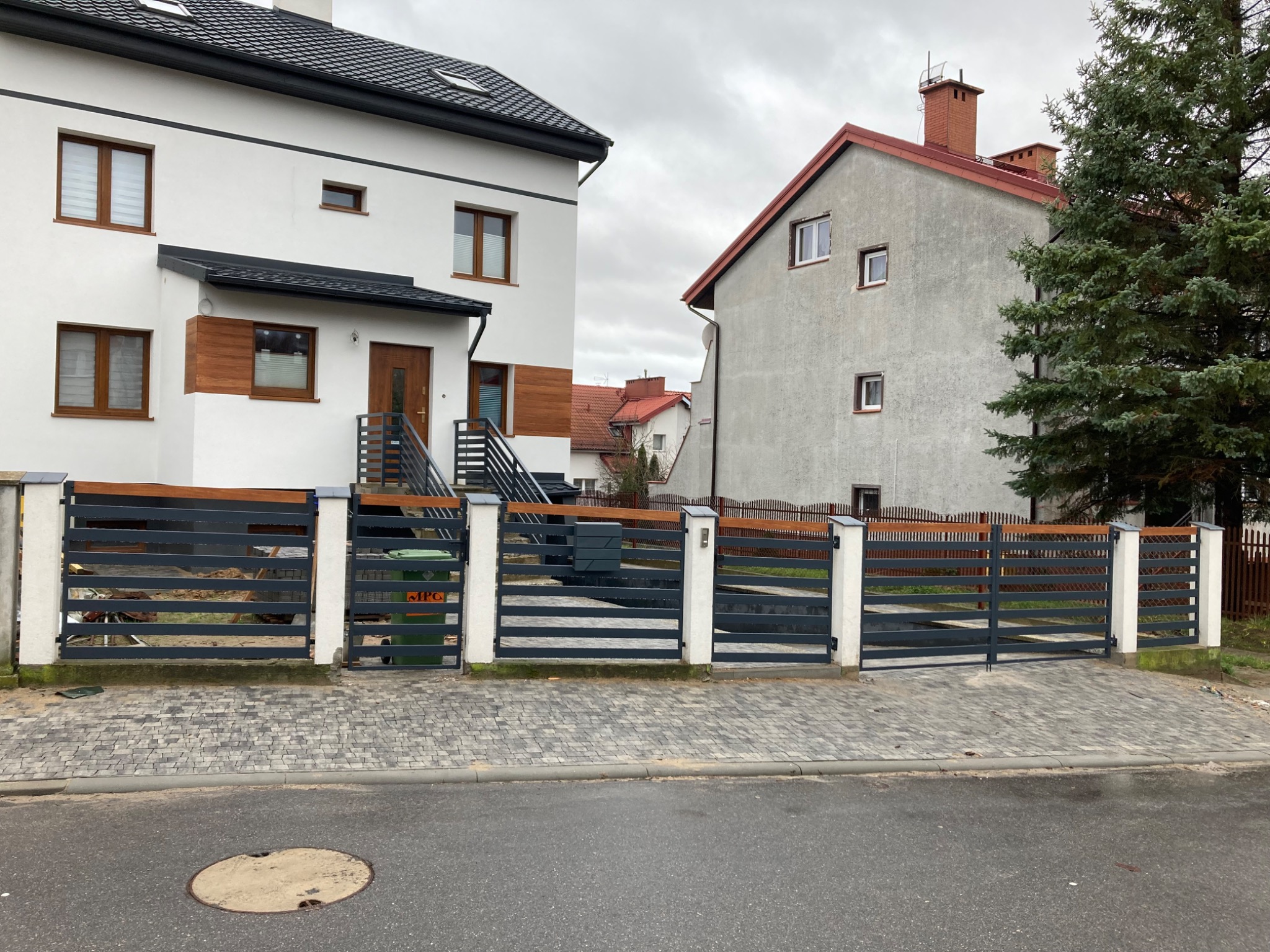 Nowoczesne ogrodzenie posesji z poziomych, grafitowych paneli i białych słupków, z drewnianym akcentem na górze. W tle dwa budynki mieszkalne i pochmurne niebo.