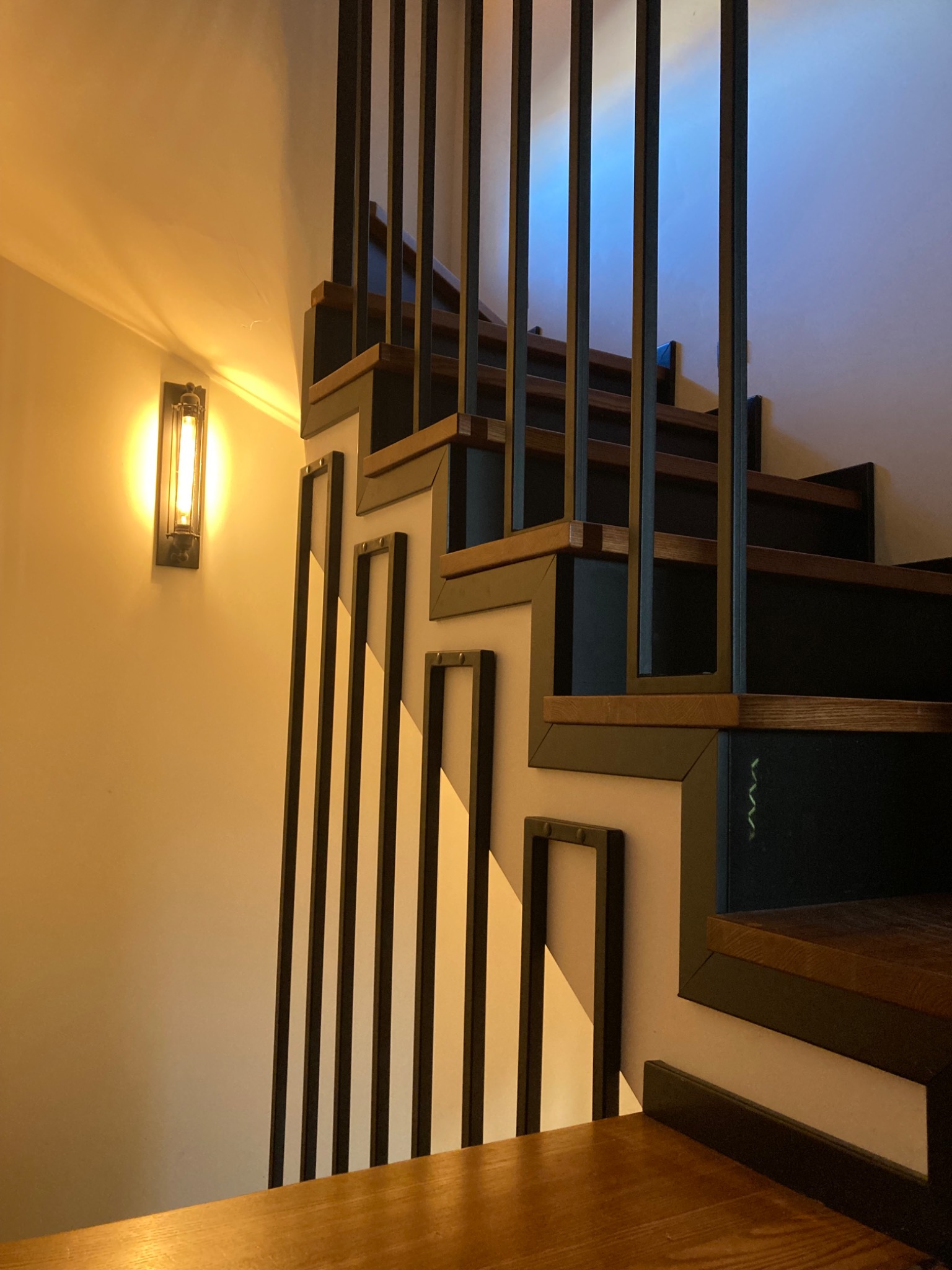 Drewniane schody z czarną, metalową balustradą o geometrycznym wzorze, oświetlone ciepłym światłem lampy ściennej. Ujęcie z dołu, ukazujące detale konstrukcji.