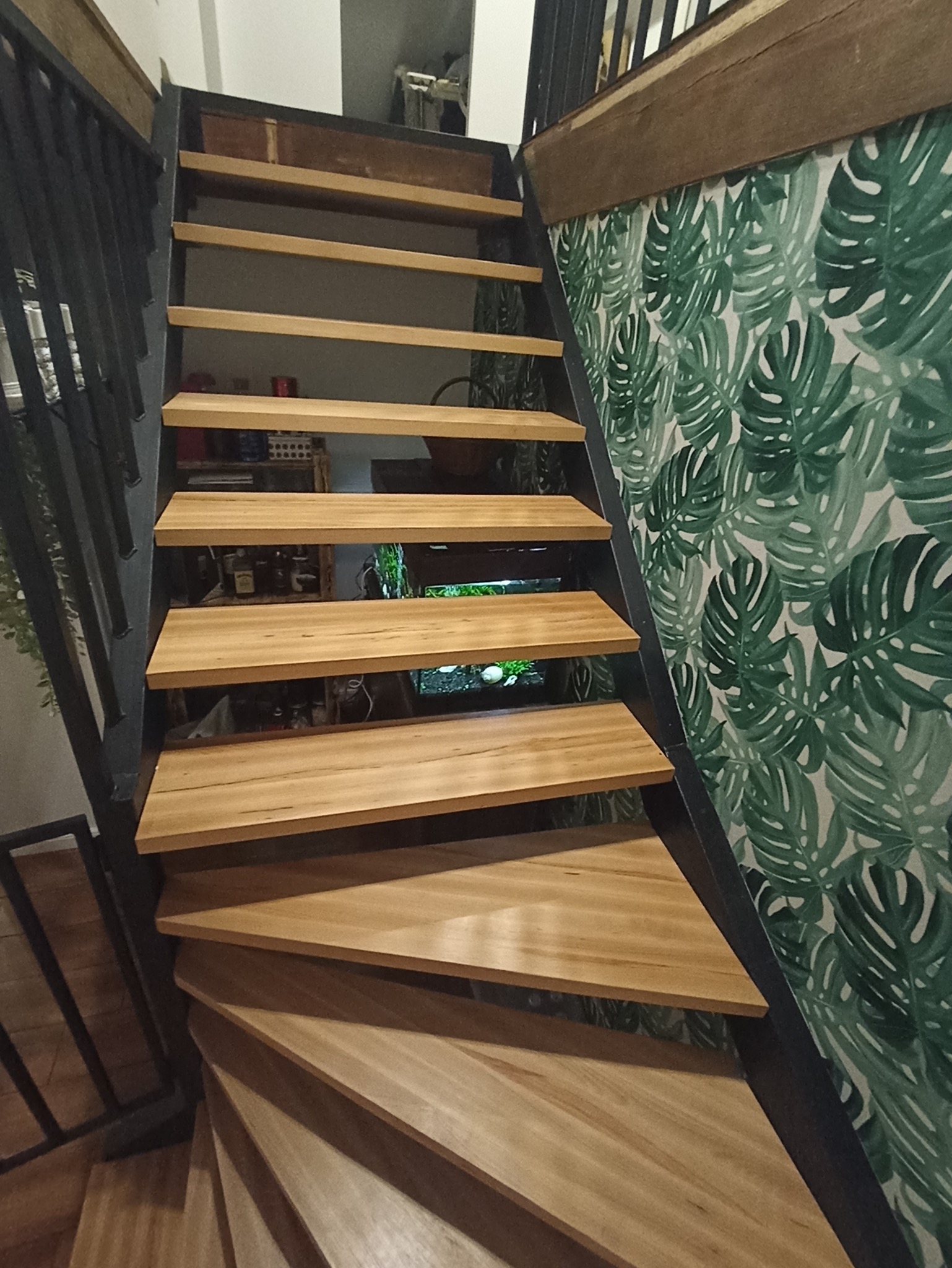 Drewniane schody z metalową balustradą, widok z dołu. Na ścianie tapeta w liście monstery. Pod schodami widoczny akwarium. Nietypowe rozwiązanie aranżacyjne.