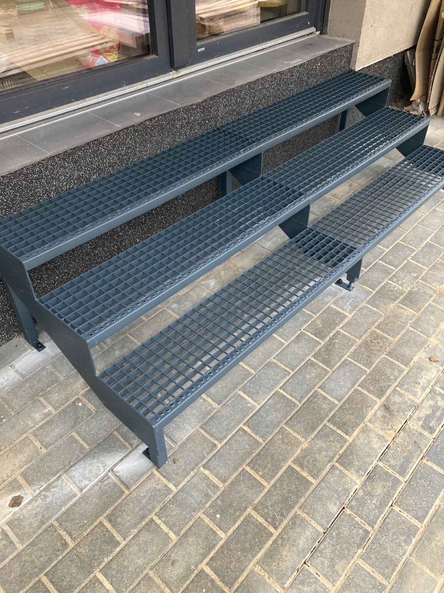 Metalowe schody zewnętrzne z kratką, prowadzące do wejścia budynku. Szare, proste stopnie na tle kostki brukowej i elewacji. Widok z góry, lekko z boku.