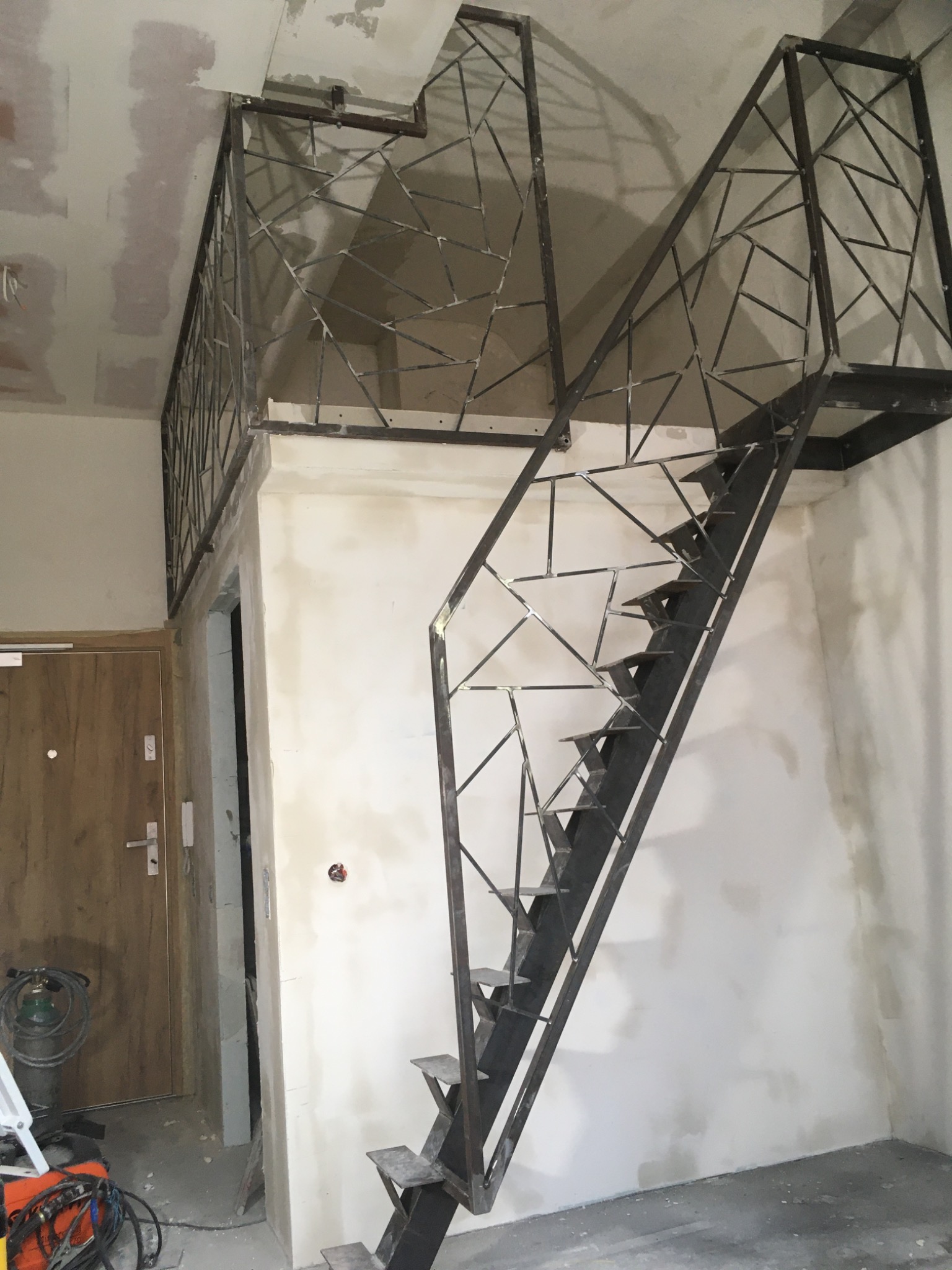 Metalowe schody z geometryczną balustradą w surowym wnętrzu. Widoczne ślady prac wykończeniowych na ścianach i suficie. W tle drzwi i sprzęt spawalniczy.