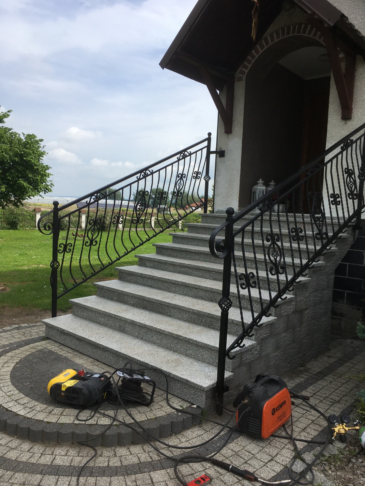 Zewnętrzne schody z szarego granitu z kutą, czarną balustradą. Na dole leżą narzędzia spawalnicze, co sugeruje niedawny montaż. Tło to zielony trawnik i błękitne niebo.