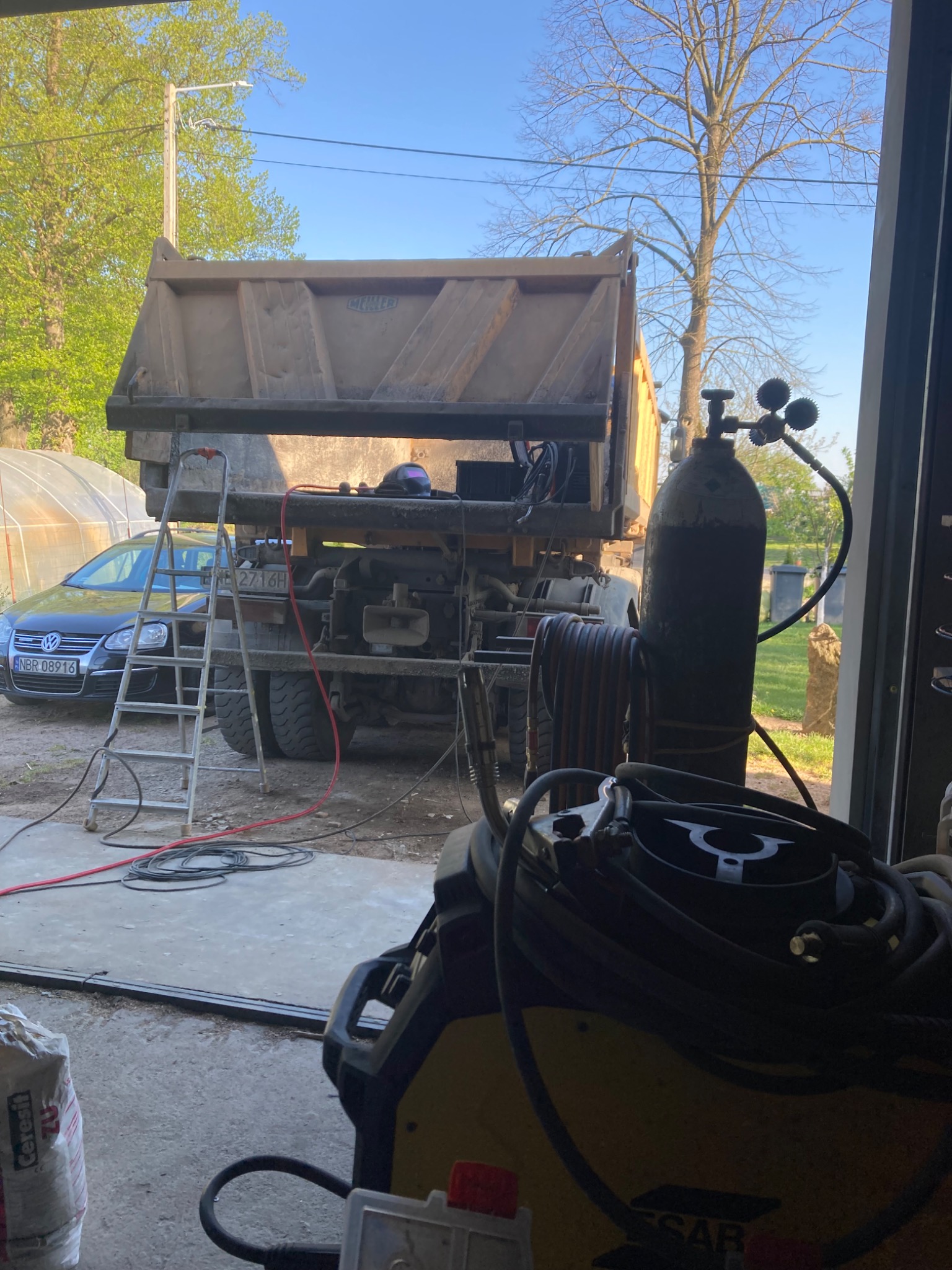 Ciężarówka wywrotka w warsztacie, widoczna drabina, butla z gazem i spawarka. W tle zaparkowany samochód osobowy i szklarnia.