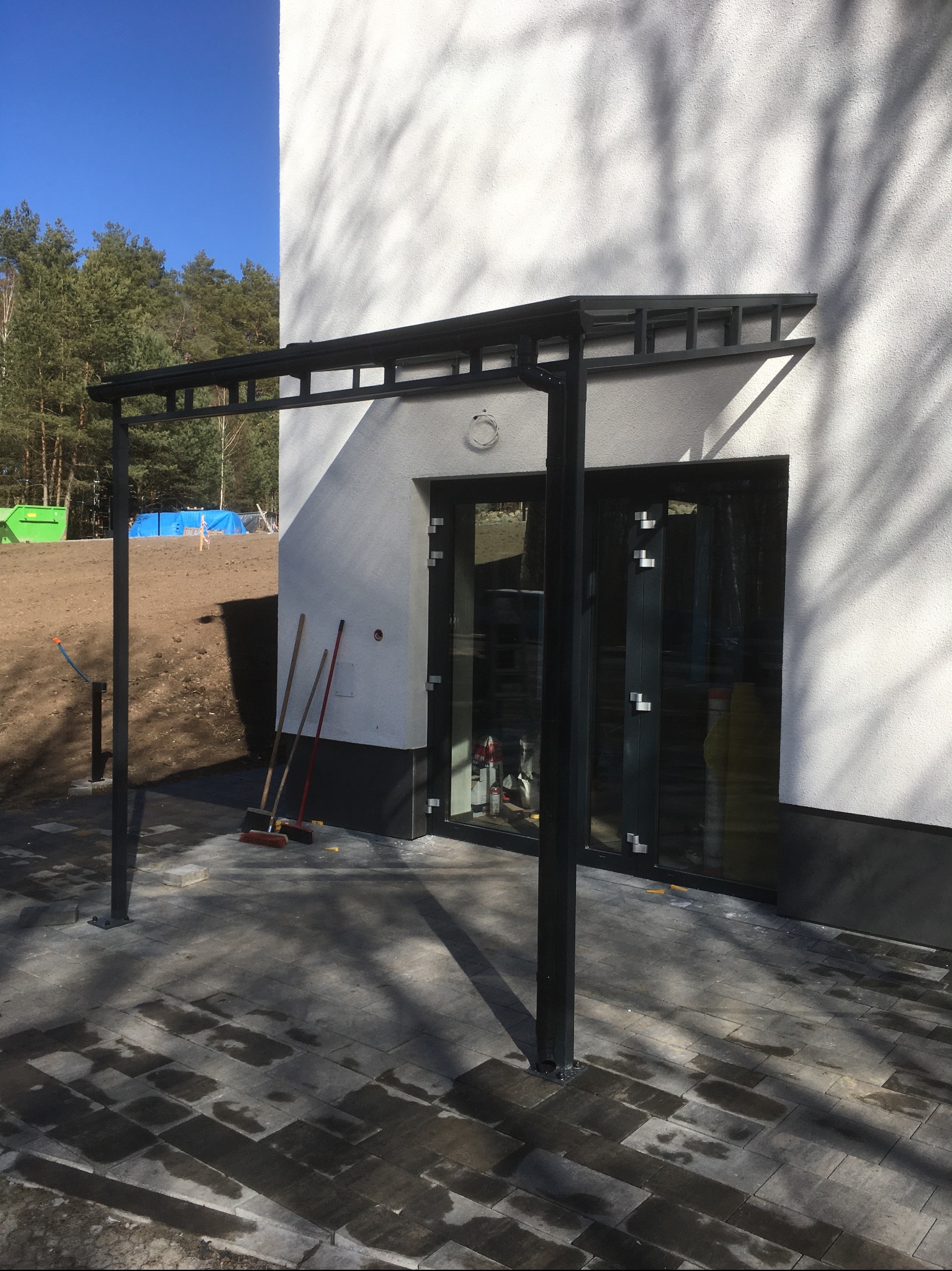 Nowoczesna, czarna pergola metalowa przy wejściu do budynku z przeszklonymi drzwiami. Widoczne narzędzia i kostka brukowa. Jasne tło z drzewami i błękitne niebo.