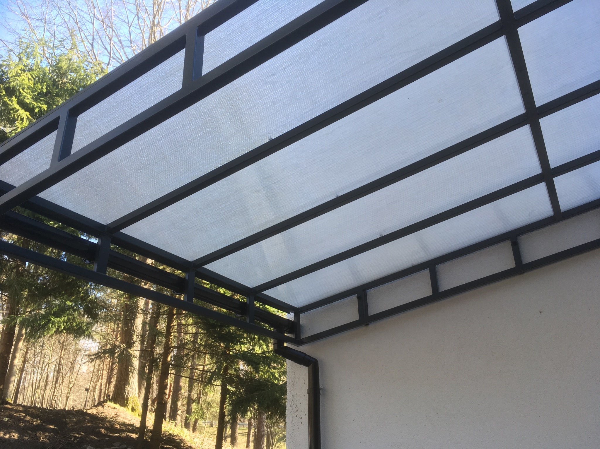 Metalowa konstrukcja z przezroczystym zadaszeniem, widok z dołu na tle drzew i fragmentu ściany budynku. Ciemna rama i jasne wypełnienie dachu.