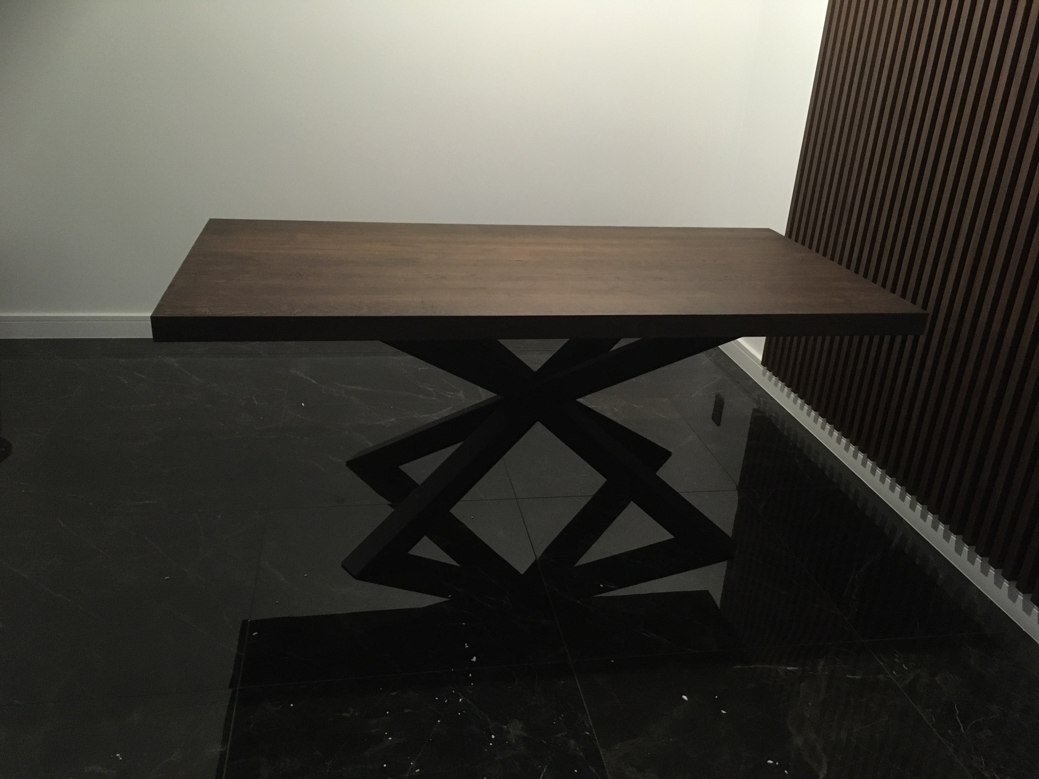 Stół z ciemnego drewna na metalowej, geometrycznej podstawie. Podłoga z czarnego marmuru, ściana biała i z brązowych paneli. Nowoczesny design.