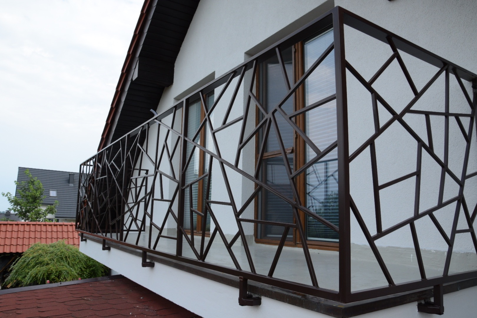 Balkon z nowoczesną, geometryczną balustradą z brązowego metalu. Widok z dołu, na tle białej ściany budynku i okien. Nietypowy wzór balustrady.