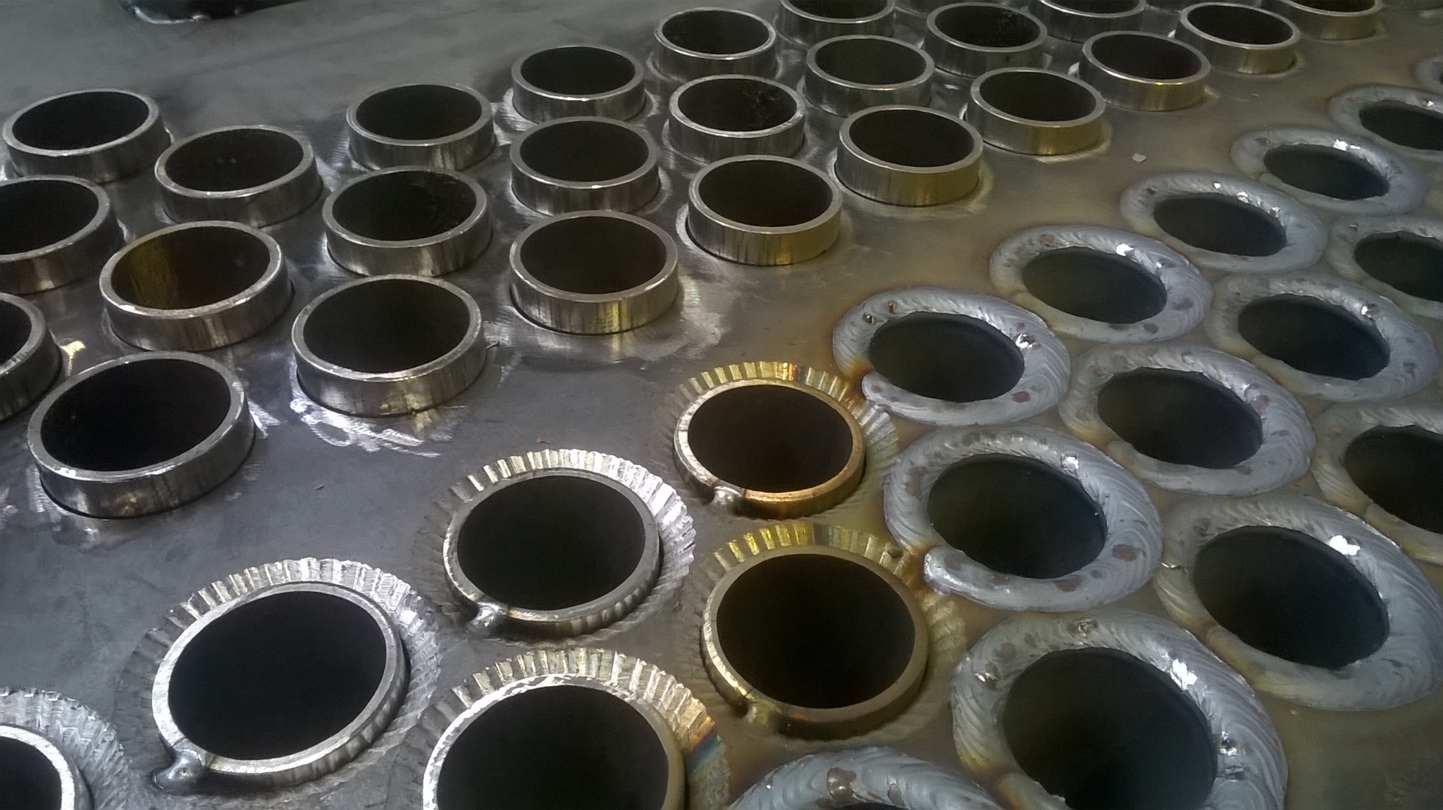 Metalowa płyta z wyciętymi otworami, część z nich z gładkimi krawędziami, a część z ząbkowanymi, widoczne ślady obróbki metalu i spawania.