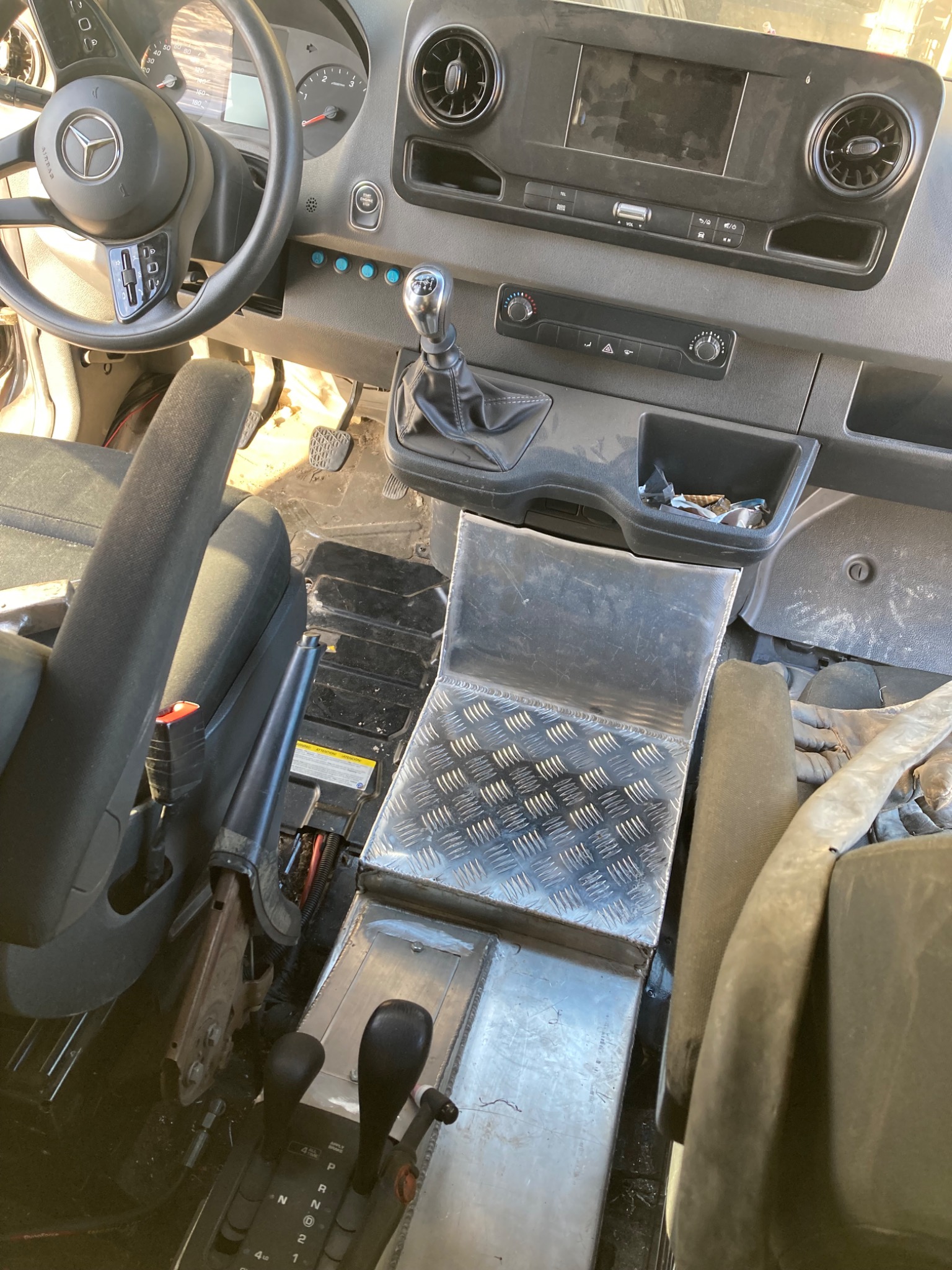 Wnętrze kabiny pojazdu dostawczego Mercedes-Benz z widoczną deską rozdzielczą, kierownicą, siedzeniami i dźwignią zmiany biegów. Widoczne elementy metalowe.