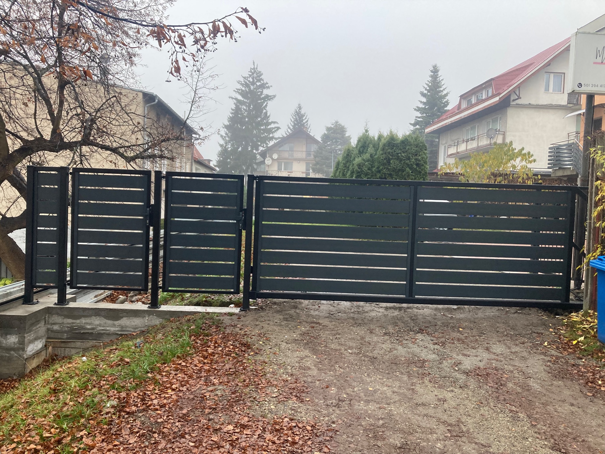 Nowoczesna, przesuwna brama w kolorze antracytowym z poziomymi panelami, zamontowana na posesji z opadłymi liśćmi. Widok na domy w tle, pochmurny dzień.