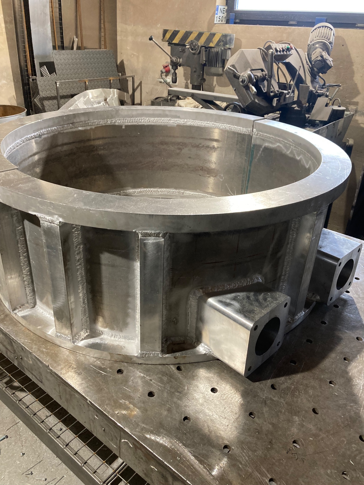 Metalowa konstrukcja cylindryczna z widocznymi spawami, umieszczona na perforowanym stole warsztatowym. W tle maszyny do obróbki metalu i tablica rejestracyjna.