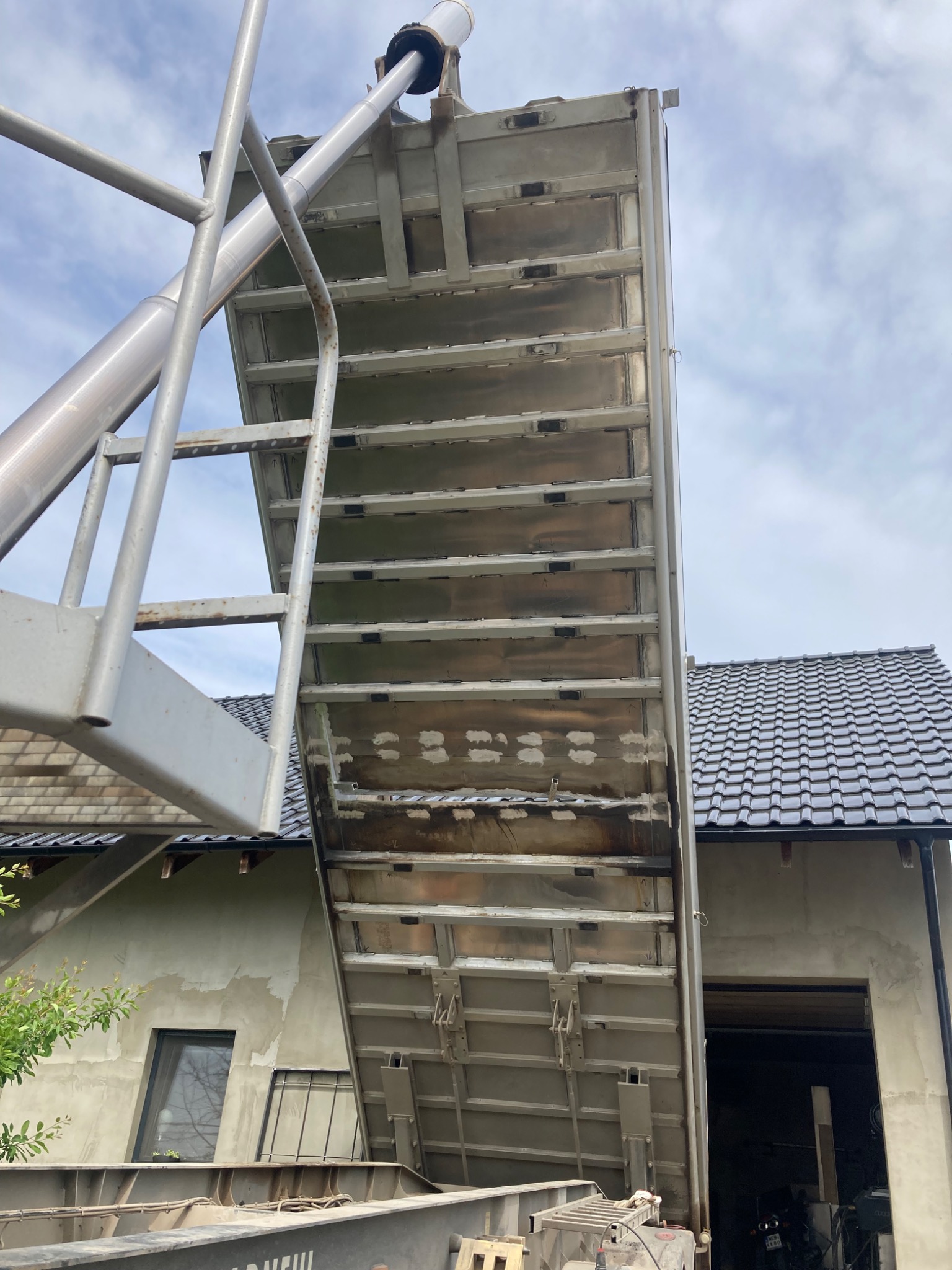 Podniesiona, metalowa platforma robocza z drabinką, widok z dołu na tle budynku z szarą elewacją i fragmentem dachu. Elementy konstrukcyjne platformy.