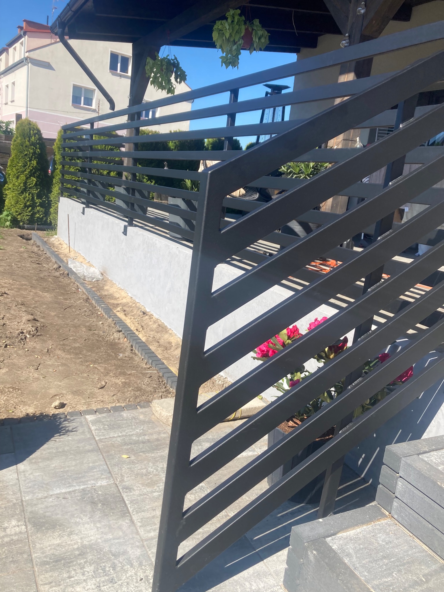 Nowoczesna, antracytowa balustrada balkonowa z poziomych profili, z widocznymi kwiatami w donicy i fragmentem tarasu z szarych płyt. Ujęcie z dołu, w słoneczny dzień.