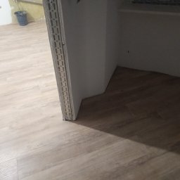 Ariel Sanetra - Jasne panele podłogowe ułożone w pokoju, widoczny fragment ściany z profilem wykończeniowym. W tle wiadro i fragment ściany w stanie surowym.