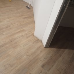 Ariel Sanetra - Ukończona podłoga z paneli w odcieniach brązu, świeżo po montażu, widoczna przy ścianie i futrynie drzwi, z narzędziami w tle.