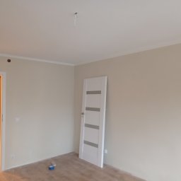 Ariel Sanetra - Jasne wnętrze po malowaniu ścian na beżowo, z białą stolarką drzwiową i listwami przypodłogowymi. Widoczny fragment drabiny malarskiej i taśma malarska.