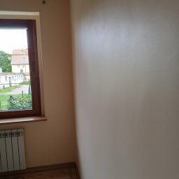 Ariel Sanetra - Jasna ściana po malowaniu, widok z rogu pokoju z oknem. Na podłodze panele i wiertarka. Grzejnik pod oknem. Świeżo pomalowane ściany w odcieniach beżu.