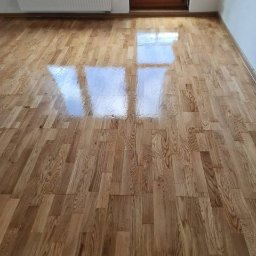 Ariel Sanetra - Parkiet dębowy ułożony w jodełkę, wykończony lakierem z wysokim połyskiem, odbijający światło z okna. Widoczny fragment grzejnika i drzwi.