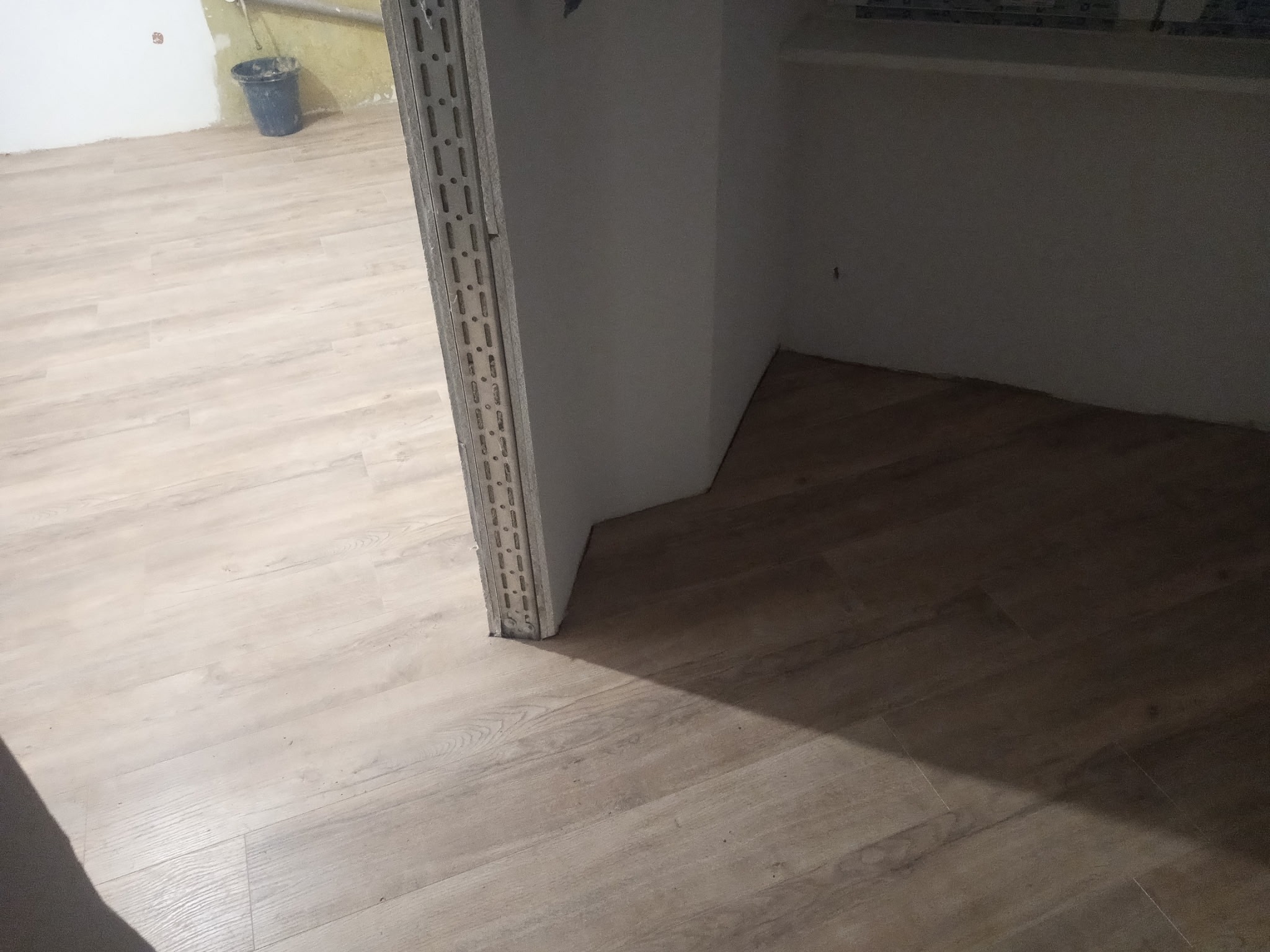 Jasne panele podłogowe ułożone w pokoju, widoczny fragment ściany z profilem wykończeniowym. W tle wiadro i fragment ściany w stanie surowym.