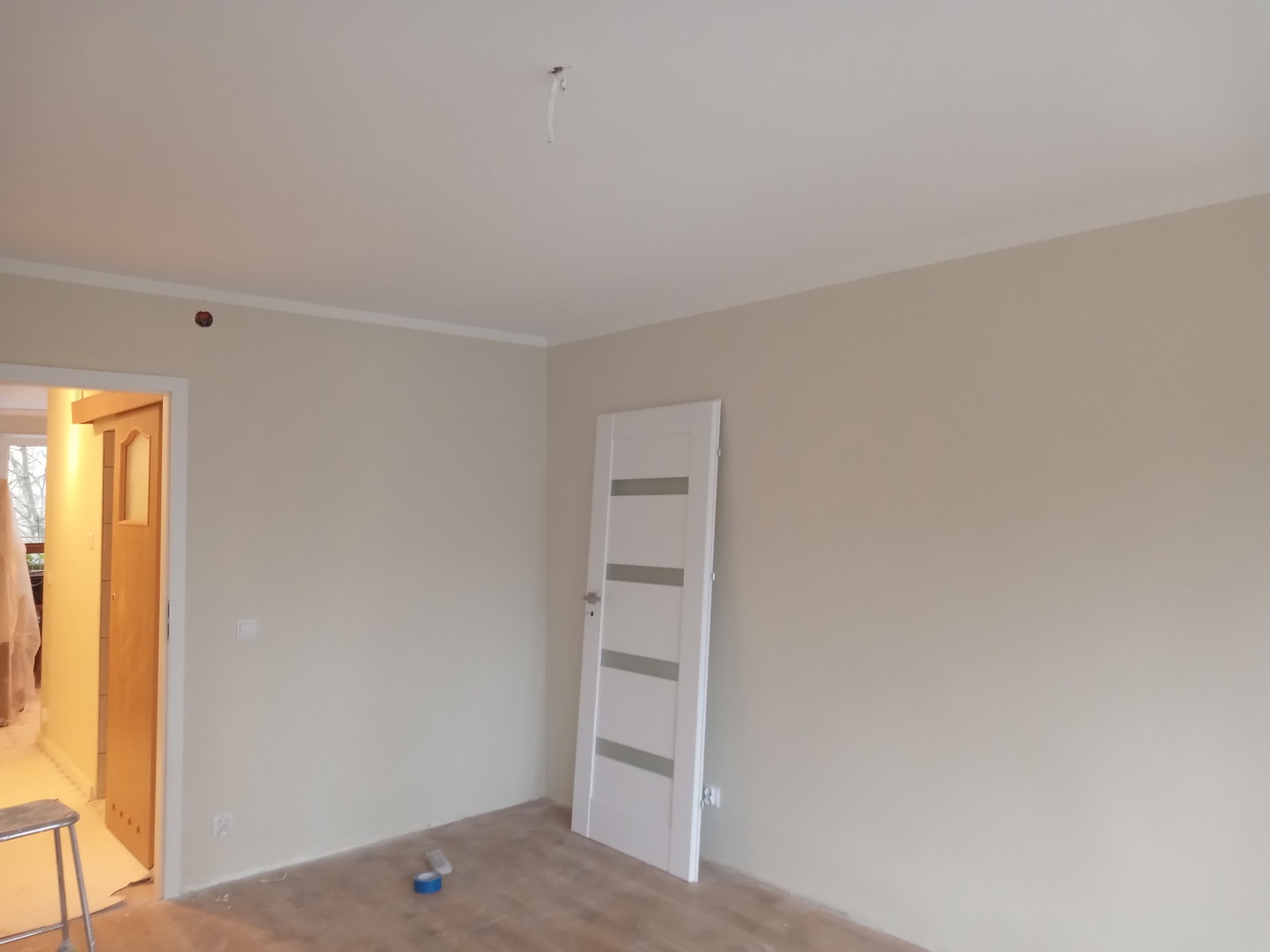 Jasne wnętrze po malowaniu ścian na beżowo, z białą stolarką drzwiową i listwami przypodłogowymi. Widoczny fragment drabiny malarskiej i taśma malarska.