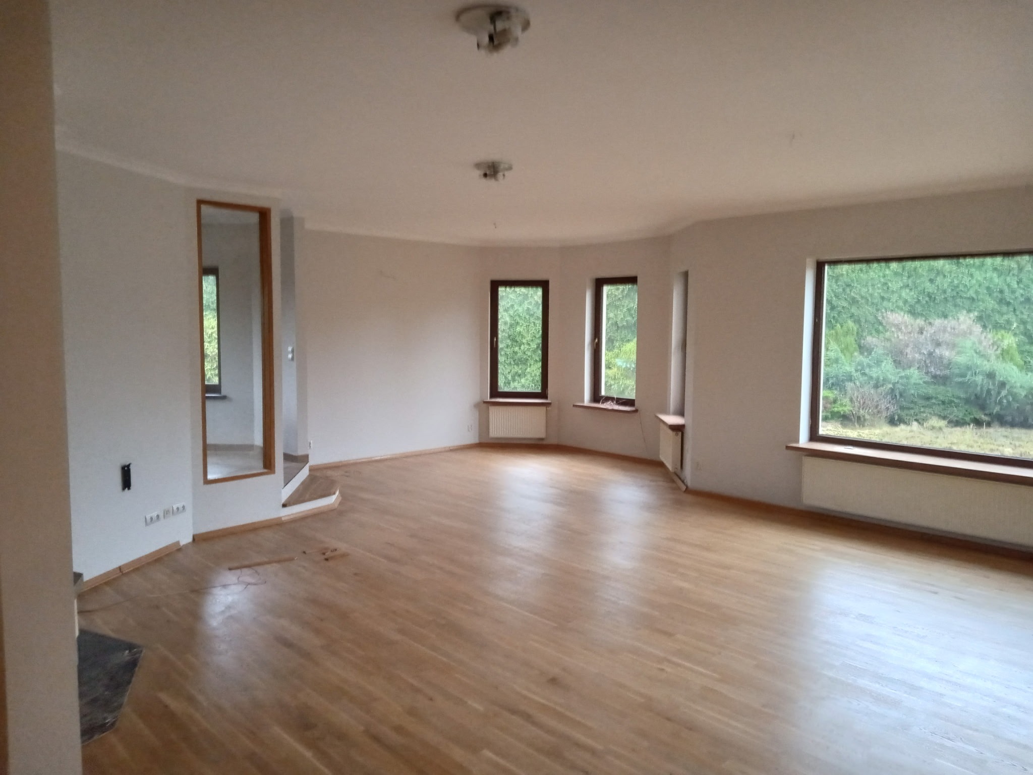 Przestronne, jasne wnętrze z parkietem i dużymi oknami z widokiem na zieleń. Ściany w odcieniach bieli, widoczne grzejniki pod oknami. Wnętrze przed wykończeniem.