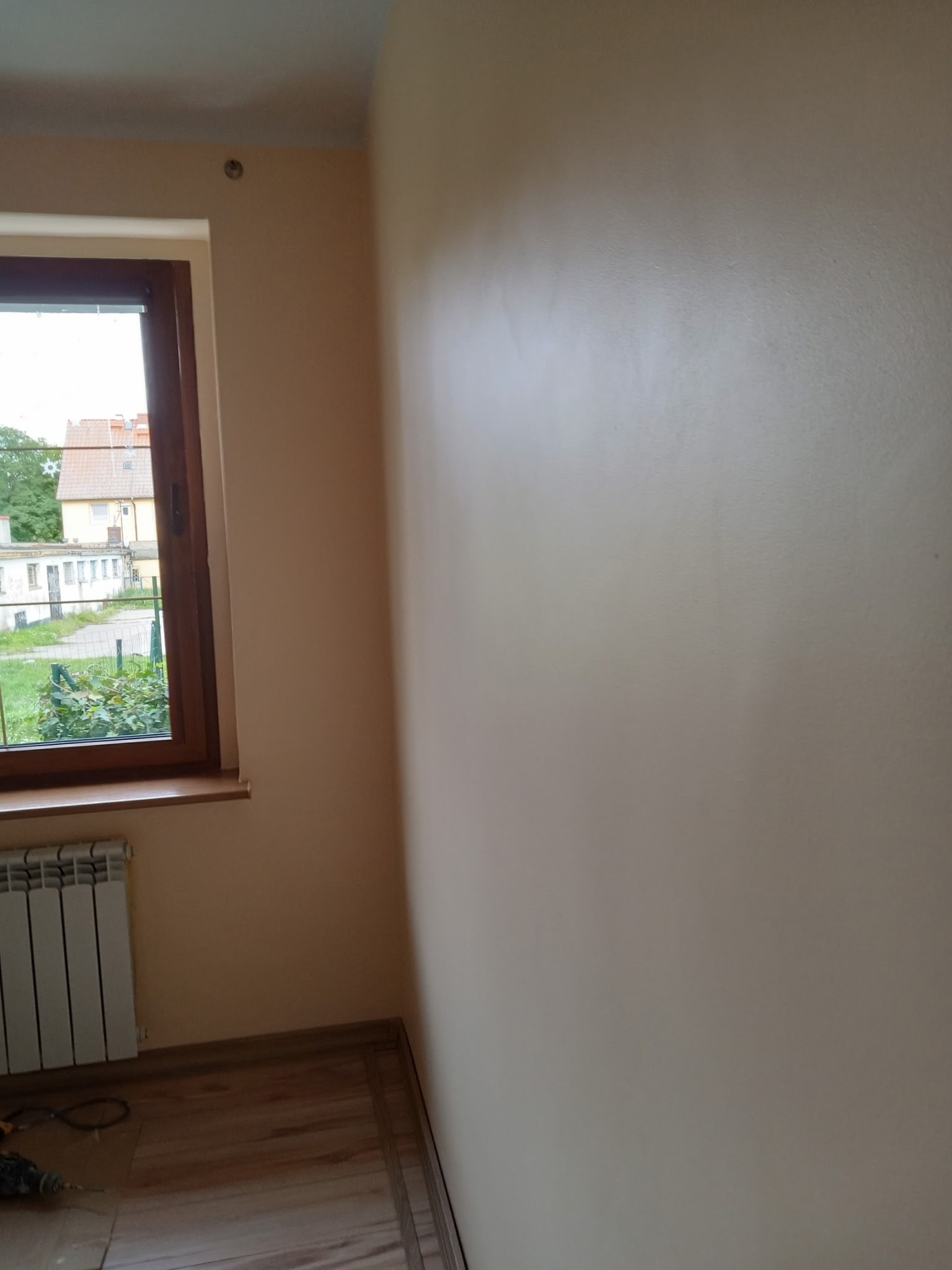 Jasna ściana po malowaniu, widok z rogu pokoju z oknem. Na podłodze panele i wiertarka. Grzejnik pod oknem. Świeżo pomalowane ściany w odcieniach beżu.