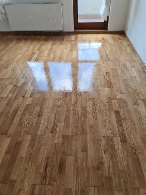 Parkiet dębowy ułożony w jodełkę, wykończony lakierem z wysokim połyskiem, odbijający światło z okna. Widoczny fragment grzejnika i drzwi.