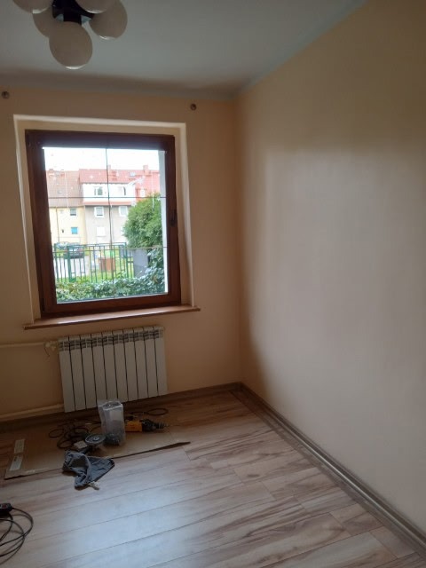 Pokój po malowaniu na beżowo z drewnianą ramą okienną. Na podłodze panele i narzędzia. Grzejnik pod oknem. Widok na budynek z zielenią za oknem.