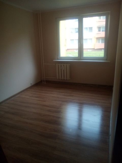 Pusty pokój po remoncie: świeżo pomalowane ściany w odcieniach beżu, parkiet z połyskiem, okno z widokiem na blok. Grzejnik pod oknem.