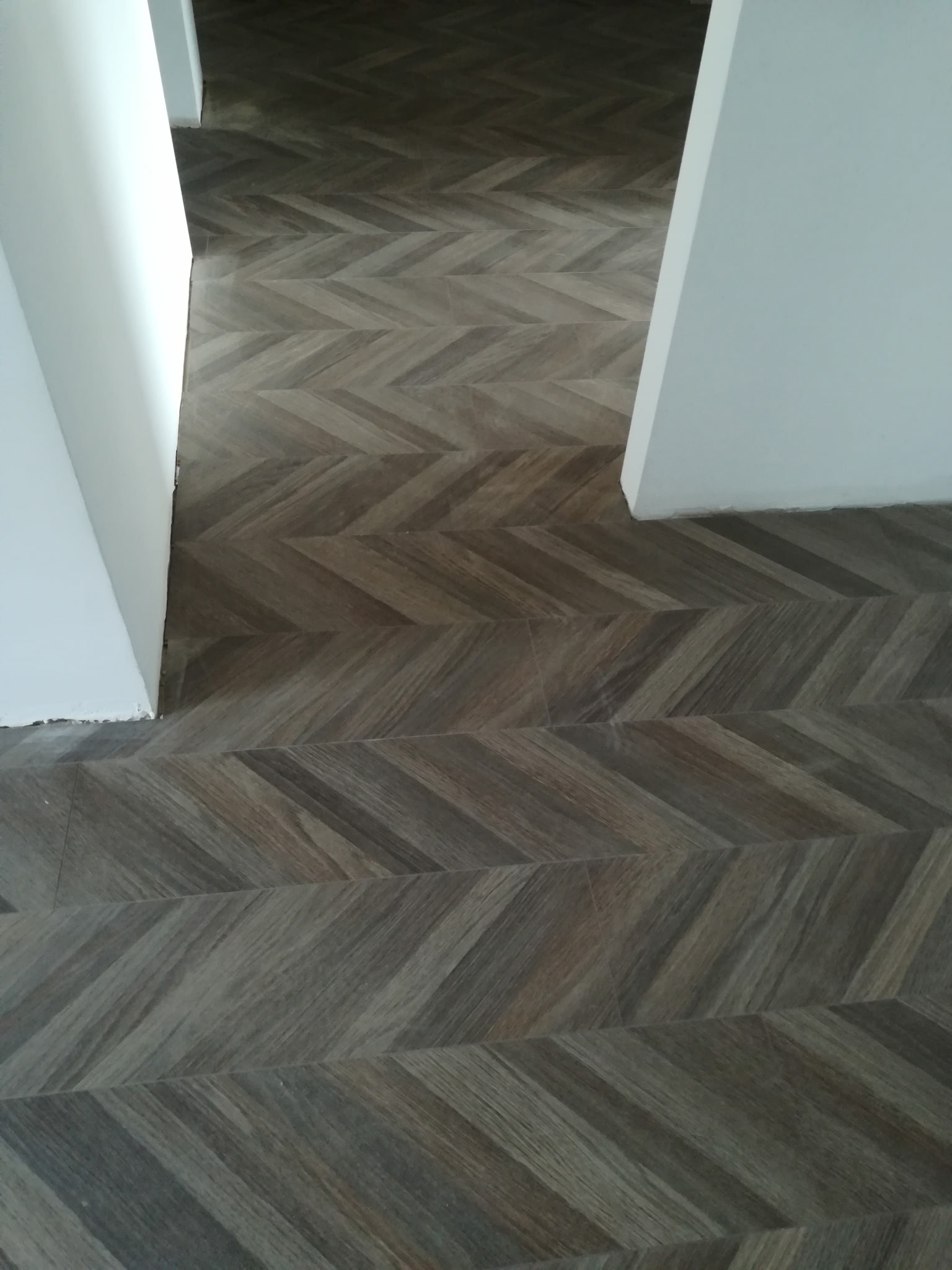 Ukończona podłoga z paneli ułożonych w jodełkę, widoczna z perspektywy podłogi, z fragmentem białej ściany po lewej stronie i przejściem do innego pomieszczenia.