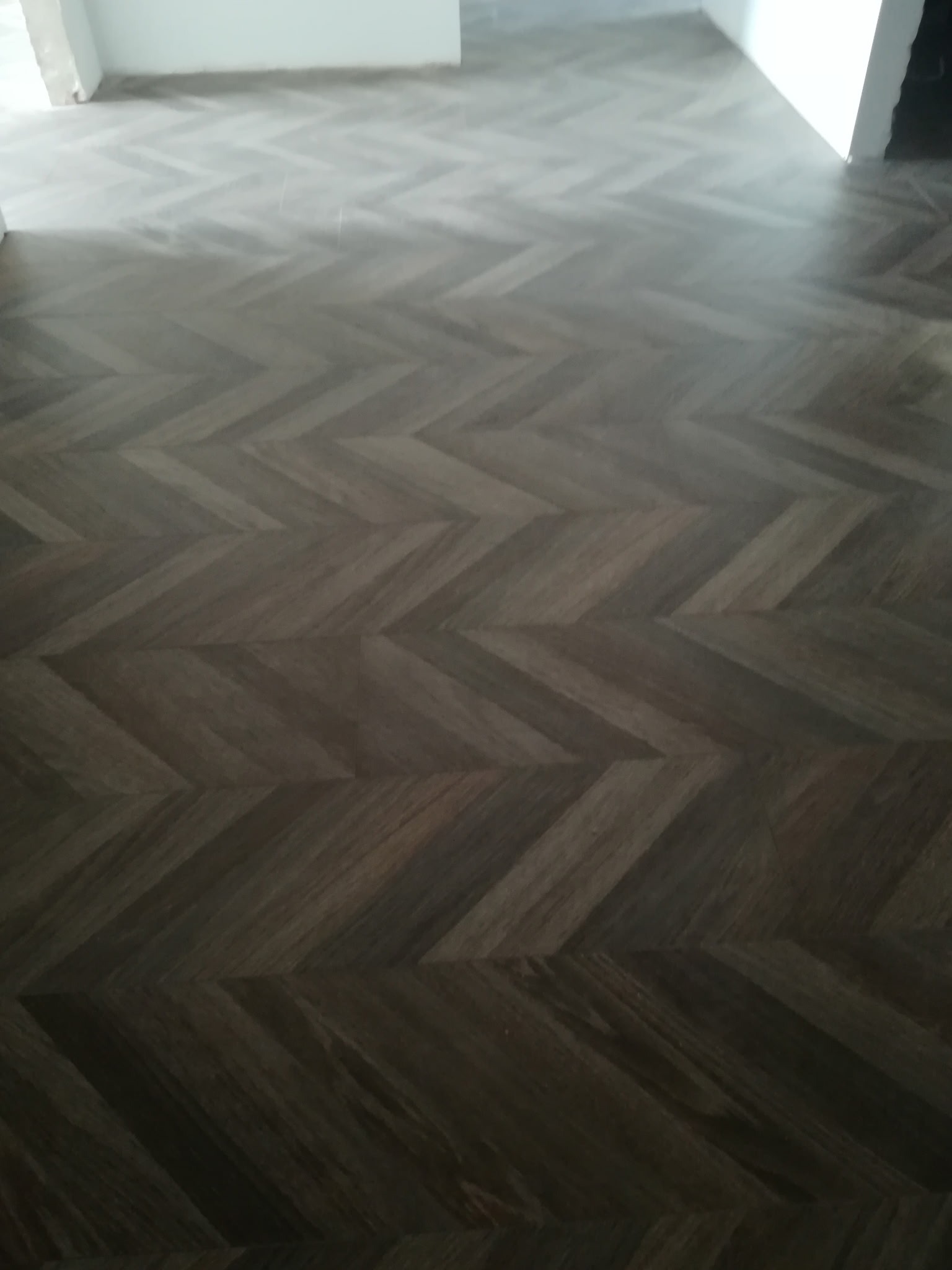 Ułożona, ciemna podłoga z paneli w jodełkę, widok z góry. W tle białe ściany i fragment otworu drzwiowego. Nowoczesny design podłogi.