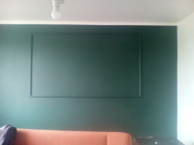 Ściana pomalowana na ciemnozielono z dekoracyjną ramą. Widoczny fragment pomarańczowej sofy i biała ściana w rogu. Minimalistyczne wnętrze po malowaniu.