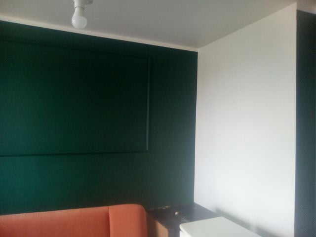 Pomieszczenie po malowaniu: ściana w odcieniach zieleni z dekoracyjną ramą i sąsiadująca biała ściana. Widoczny fragment pomarańczowej sofy i lampa sufitowa.