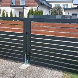 Megamet - Nowoczesna brama wjazdowa z czarnych, poziomych profili stalowych i drewnopodobnych akcentów, na tle domów jednorodzinnych i żwirowego podjazdu.