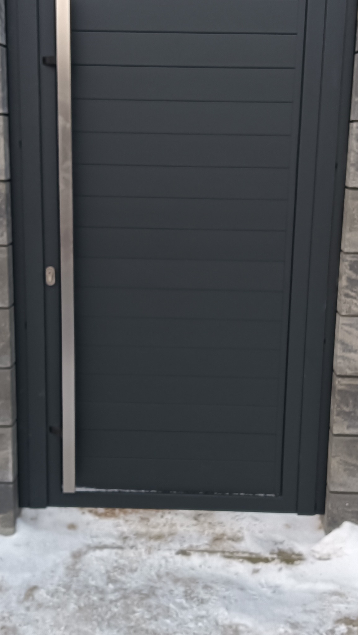 Nowoczesna, pozioma brama wjazdowa w kolorze antracytu, z widocznym srebrnym uchwytem i fragmentem słupka z szarej kostki. U dołu śnieg.