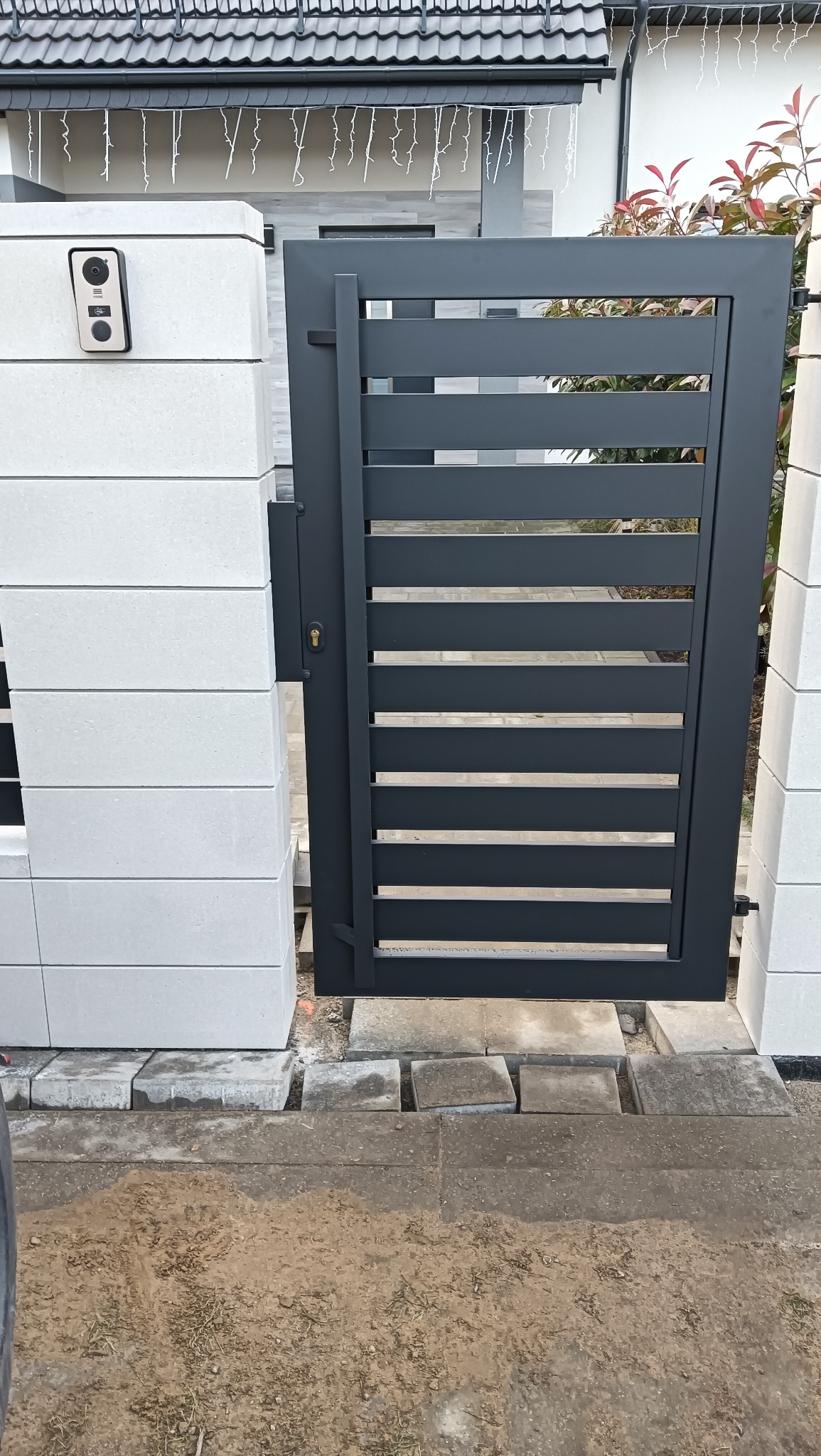 Nowoczesna, grafitowa furtka z poziomymi szczeblami, zamontowana między białymi, betonowymi słupkami. Widoczny dom z szarym dachem i dekoracyjnym oświetleniem.