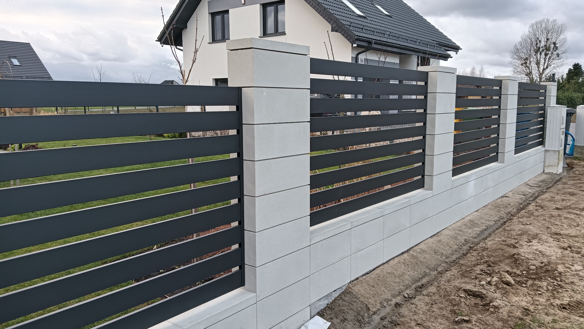 Nowoczesne ogrodzenie metalowe z poziomymi panelami w kolorze grafitowym i białymi słupkami z bloczków betonowych na tle domu jednorodzinnego.