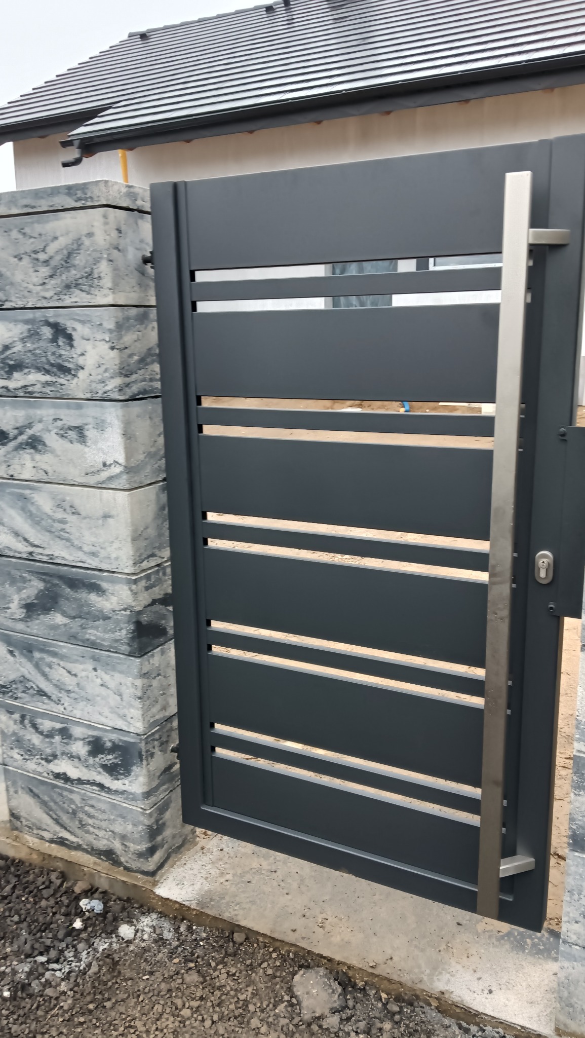 Nowoczesna, grafitowa brama wjazdowa z poziomymi szczebelkami i srebrnym uchwytem, osadzona na betonowym słupku o marmurkowym wzorze, na tle domu z szarą dachówką.