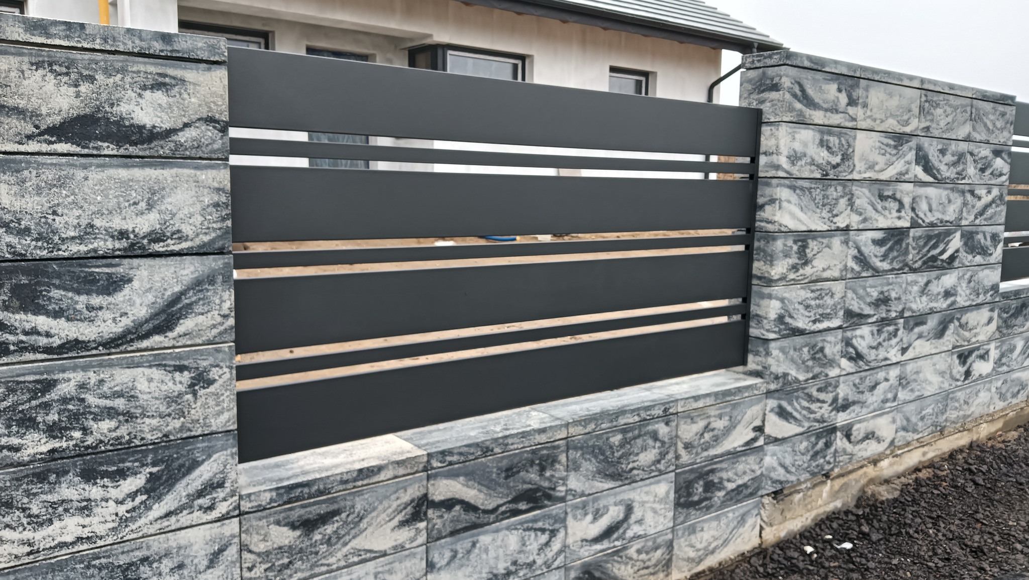 Nowoczesne ogrodzenie z szarego metalu z poziomymi prętami i kamiennymi słupkami o marmurkowym wzorze, otaczające posesję z domem w tle.