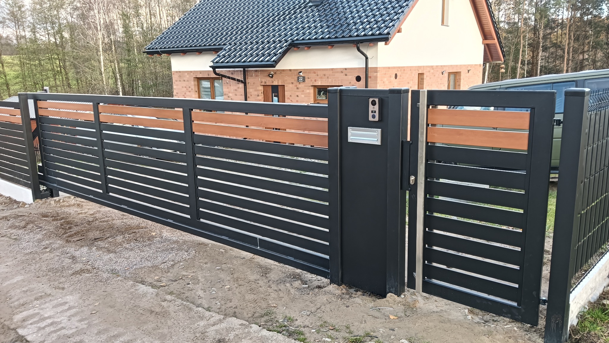 Nowoczesna brama wjazdowa przesuwna z czarnych, poziomych profili i drewnianymi akcentami, z wideodomofonem i skrzynką na listy, na tle domu z ciemną dachówką.