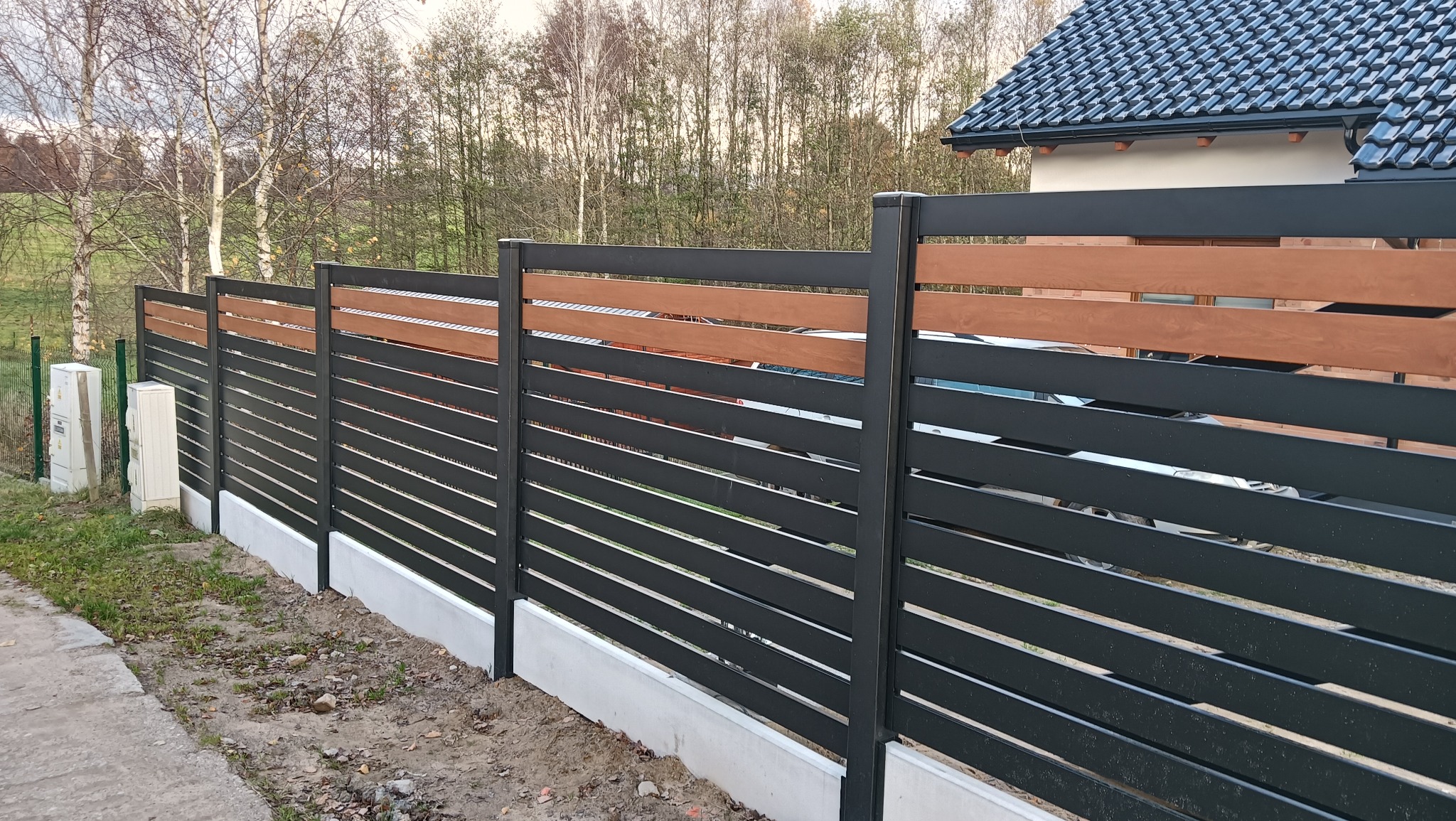 Nowoczesne ogrodzenie posesji z poziomych, czarnych paneli metalowych i drewnianych akcentów, osadzone na betonowej podmurówce. Widok z zewnątrz, w tle drzewa.