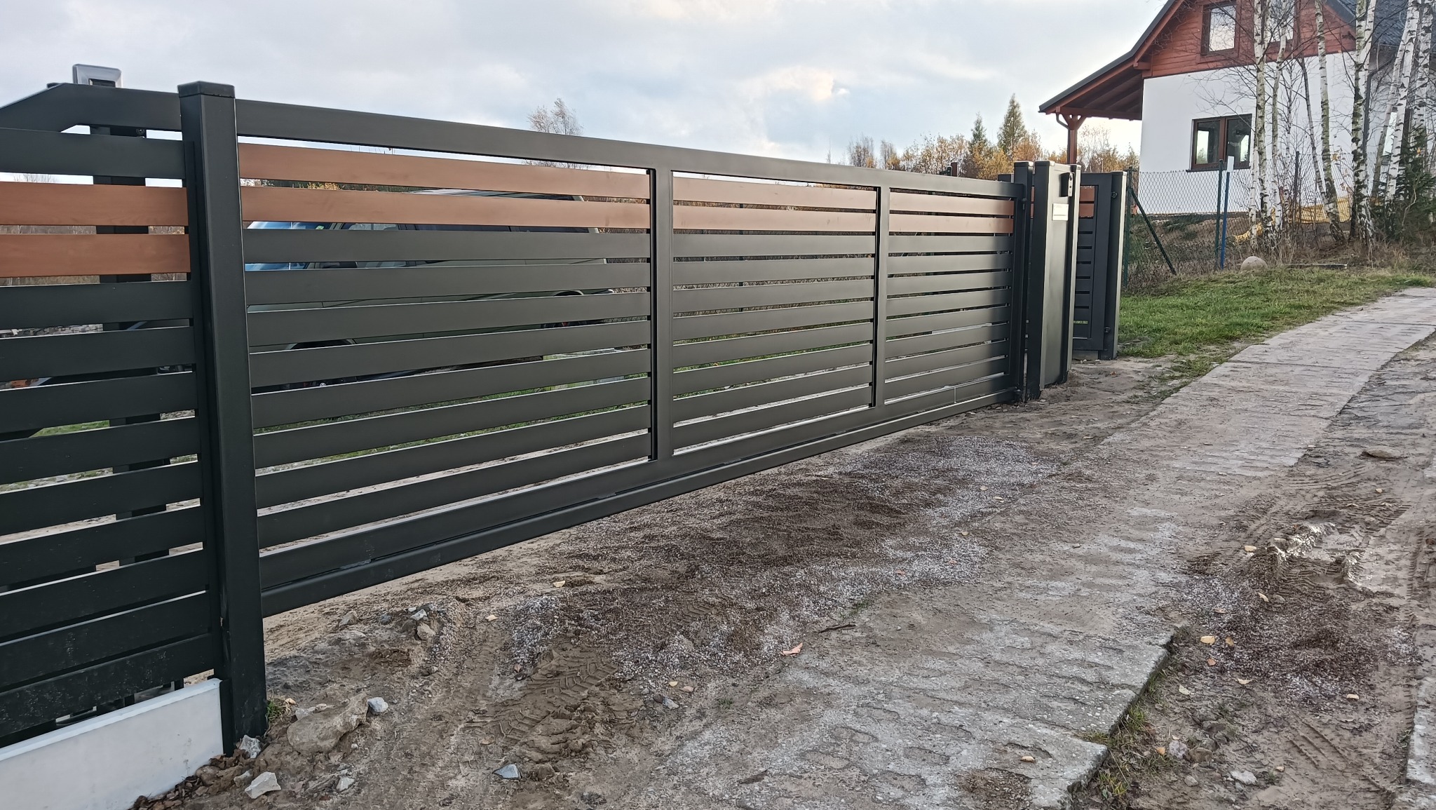 Nowoczesna, przesuwna brama wjazdowa z poziomymi, grafitowymi panelami i akcentami w kolorze drewna. Widoczny fragment domu w tle. Brama zamontowana na posesji.