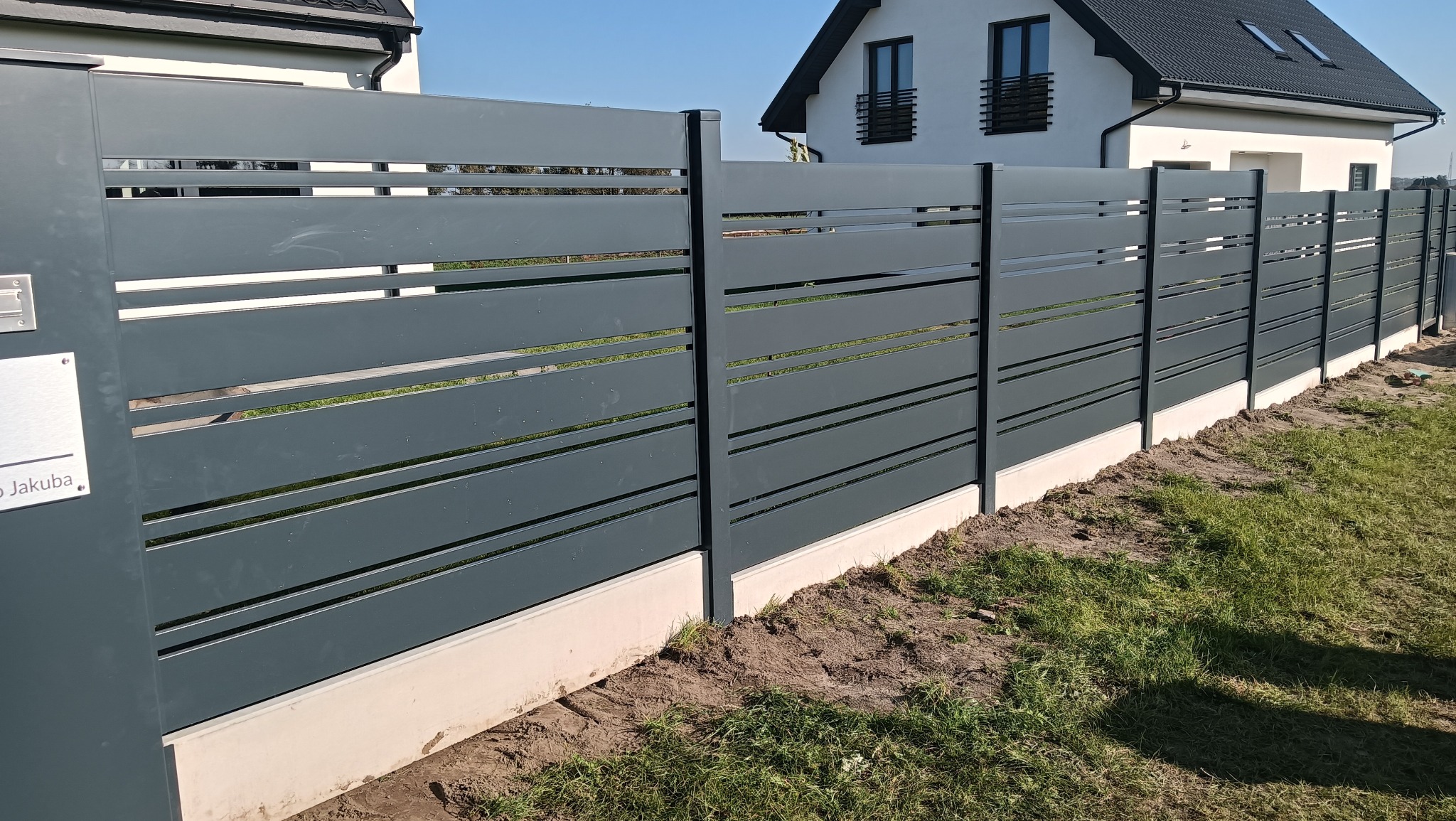 Nowoczesne, poziome ogrodzenie metalowe w kolorze grafitowym z subtelnymi prześwitami, osadzone na betonowej podmurówce. W tle dom jednorodzinny.
