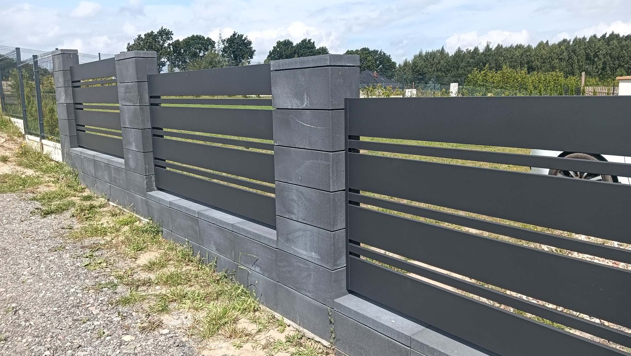 Nowoczesne ogrodzenie z poziomych, grafitowych paneli i betonowych słupków, z widocznym fragmentem siatki ogrodzeniowej w tle, na tle zieleni i błękitnego nieba.