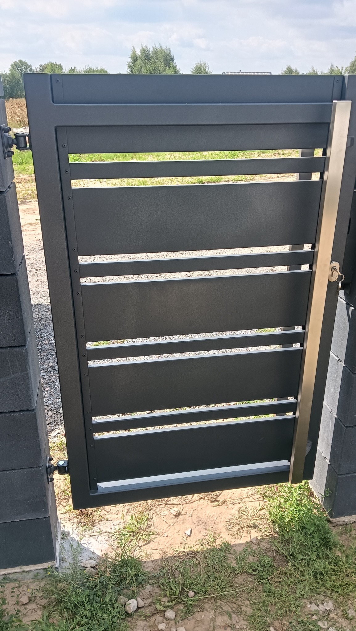 Nowoczesna, grafitowa brama wjazdowa z poziomymi panelami, zamontowana na betonowych słupkach. Widoczny zamek i klucz. Tło stanowi trawa i drzewa.