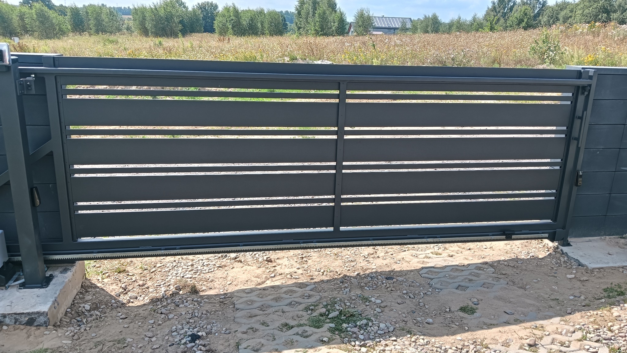 Nowoczesna, przesuwna brama wjazdowa z poziomymi panelami, szara, zamontowana na posesji z widocznym mechanizmem otwierania i zielonym tłem. Brama z bliska.