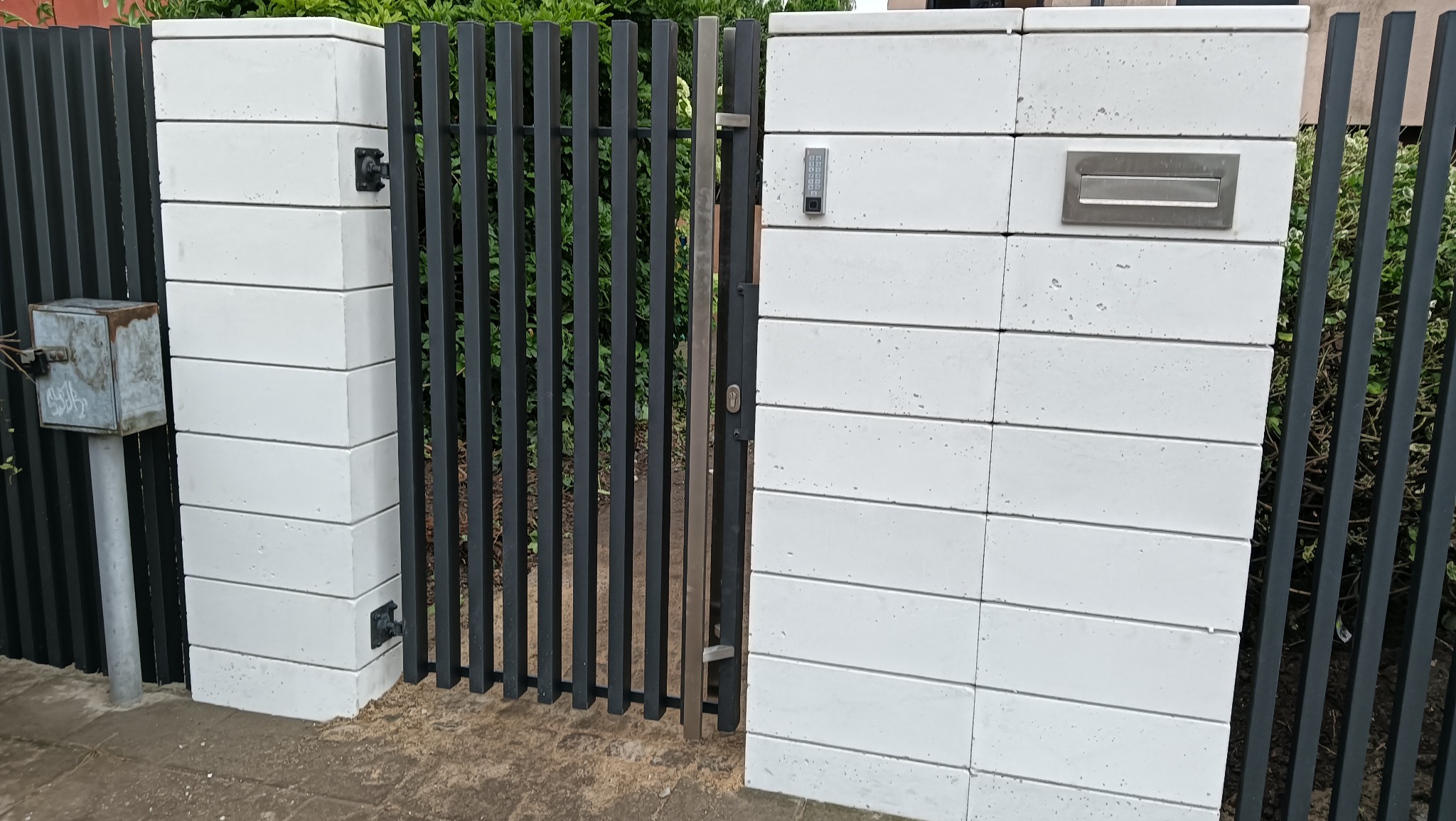 Nowoczesna brama wjazdowa z czarnymi, pionowymi prętami i białymi, poziomymi bloczkami, z zamontowaną skrzynką na listy i panelem numerycznym.