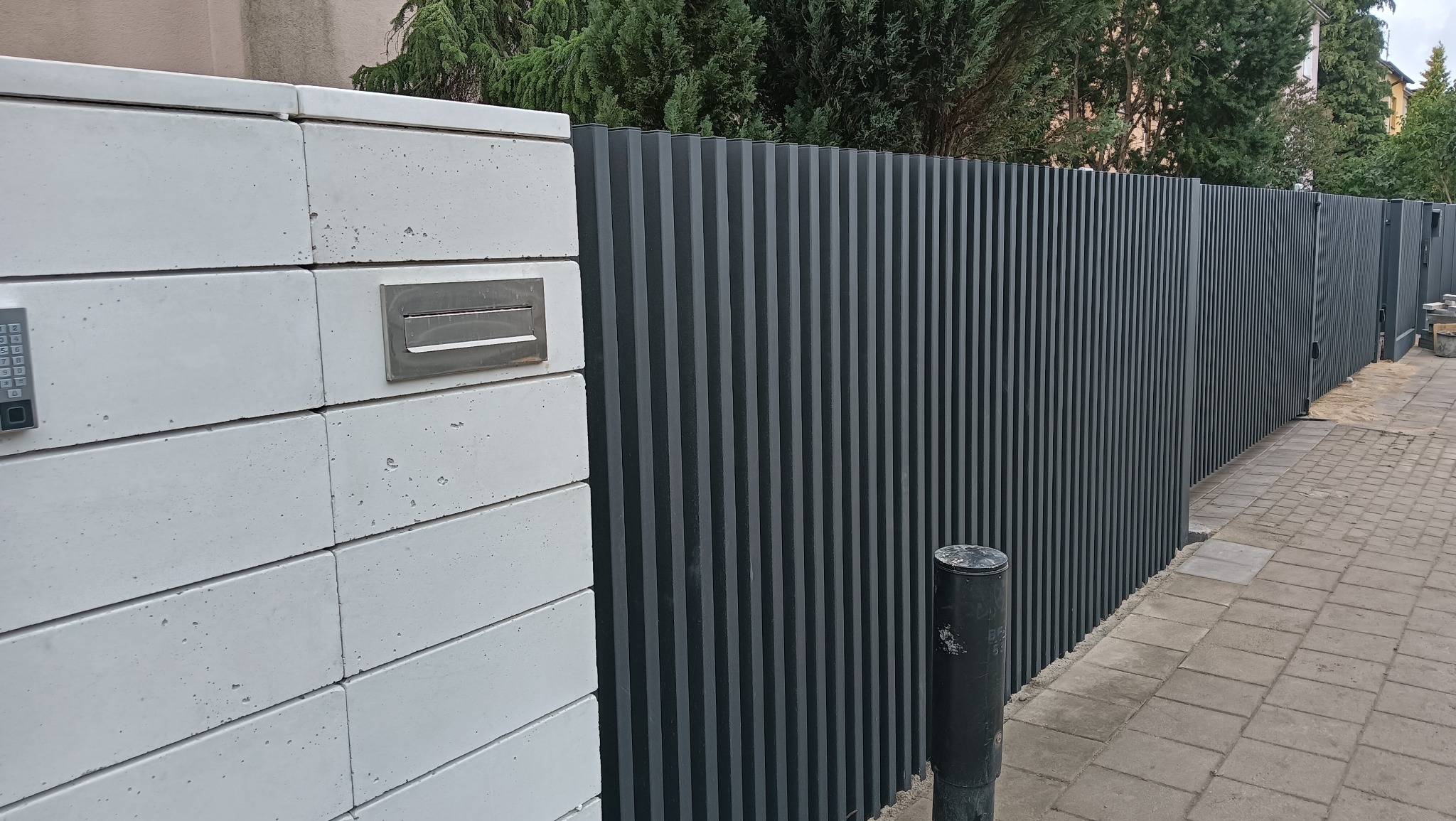 Nowoczesne ogrodzenie z szarych, pionowych paneli metalowych i białego murku z wbudowaną skrzynką na listy oraz panelem numerycznym, widok z poziomu chodnika.