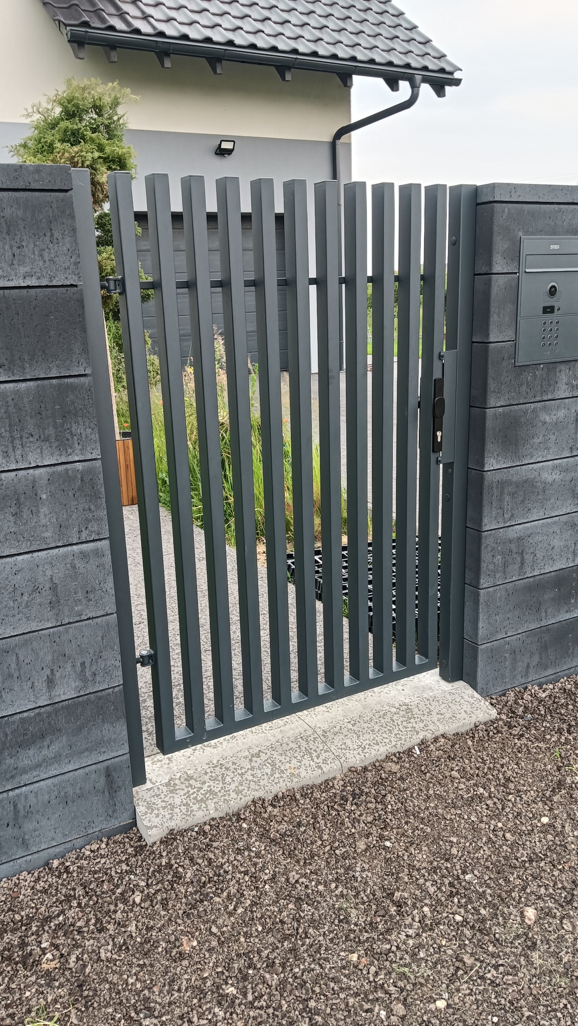 Nowoczesna, grafitowa furtka z pionowych profili, osadzona między betonowymi słupkami z domofonem. Widok na posesję przez otwartą furtkę.
