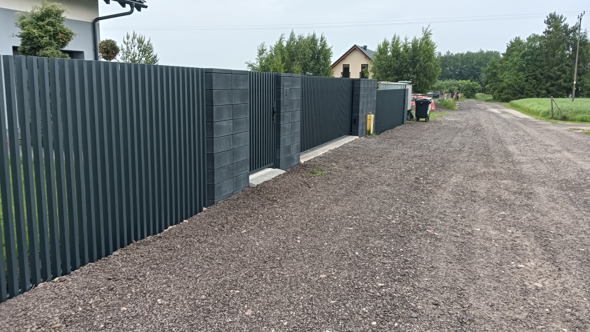 Nowoczesne, grafitowe ogrodzenie panelowe z bramą wjazdową, wykończone betonowymi słupkami. Widok na żwirową drogę i dom w tle. Minimalistyczny design.